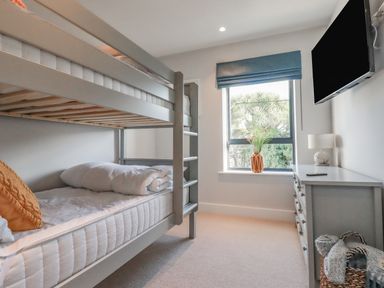 A bedroom with a bunk bed a dresser with a lamp a window with a blue blind and a TV mounted on the wall at 7 Longshore in St Columb Minor