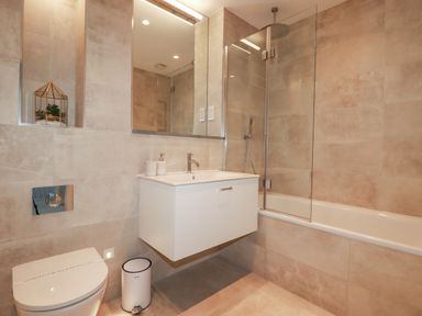 A bathroom with a toilet, floating sink cabinet, bathtub with glass shower screen, and a large mirror at 7 Longshore in St Columb Minor