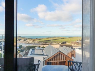 A balcony with chairs and a table overlooking rooftops sandy beach grassy hills and the sea at 7 Longshore in St Columb Minor