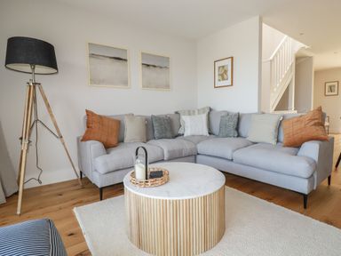 A living room with a gray sectional sofa pillows a round coffee table wooden floor and a tripod floor lamp at 7 Longshore in St Columb Minor