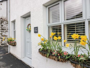 A white cottage exterior with a door and windows with flower boxes containing yellow daffodils at The Anvil in Broughton-In-Furness