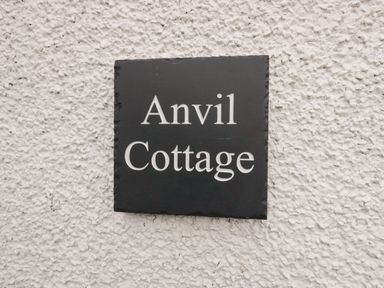A black sign with white text reading Anvil Cottage on a rough white wall at The Anvil in Broughton-In-Furness