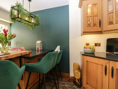 A kitchen with wooden cabinets green chairs a fruit bowl flowers and a wooden rooster figurine at The Anvil in Broughton-In-Furness