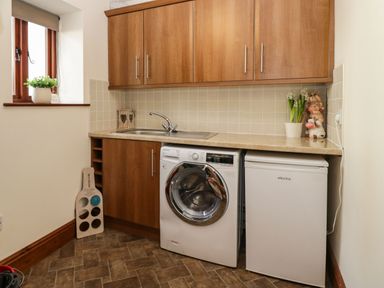 A utility room with wooden cabinets a washing machine a small fridge a sink and plants on the countertop at The Anvil in Broughton-In-Furness