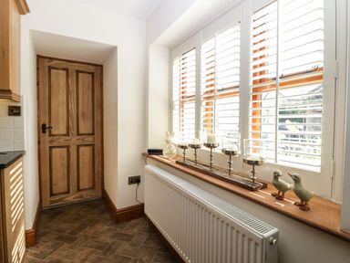 A hallway with a wooden door on the left and a window with white shutters and decorative items on the windowsill including candles and figurines at The Anvil in Broughton-In-Furness