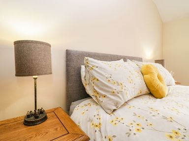 A bedroom with a bed with floral bedding and a yellow round pillow next to a wooden nightstand with a lamp at The Anvil in Broughton-In-Furness