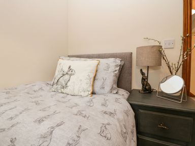 A bedroom with a bed covered in rabbit printed bedding and a pillow next to a bedside table with a lamp a rabbit figurine and a round mirror at The Anvil in Broughton-In-Furness