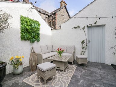 An outdoor patio with a corner sofa a table with flowers and two stools at The Anvil in Broughton-In-Furness