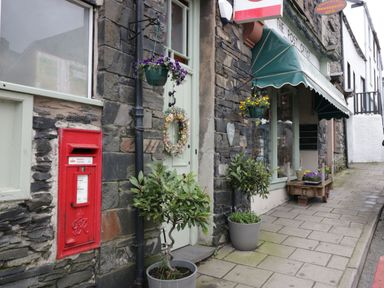 Outside view of a post office with stone walls a red postbox potted plants hanging flower baskets and a green awning at The Anvil in Broughton-In-Furness