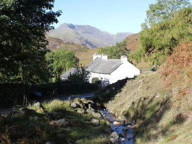 A small stream flows beside a white house with trees and mountains in the background at The Anvil in Broughton-In-Furness