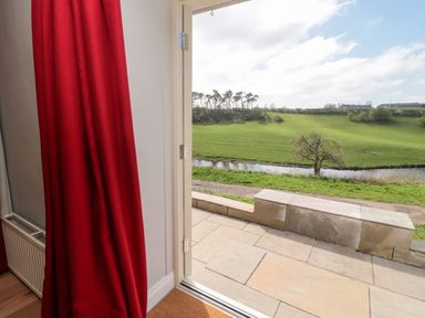 An open door with a red curtain looking out to a patio and green field with a tree and a small water body at Whiteadder Bank in Berwick-Upon-Tweed