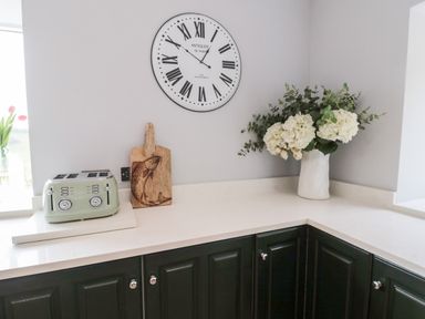 A kitchen corner with a toaster a wooden cutting board with a fish illustration a large wall clock and a white vase with white flowers at Whiteadder Bank in Berwick-Upon-Tweed