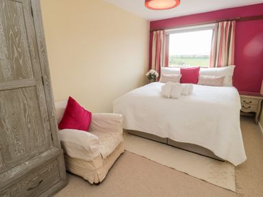 A bedroom with a double bed with white bedding and pillows a beige armchair with a red cushion a wooden wardrobe and a window with curtains at Whiteadder Bank in Berwick-Upon-Tweed