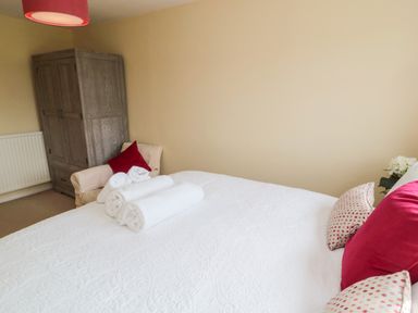 A bedroom with a white bedspread red cushions rolled towels an armchair a wooden wardrobe and a red lampshade at Whiteadder Bank in Berwick-Upon-Tweed