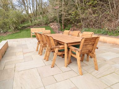 A wooden outdoor dining table with eight chairs on a stone patio and a wooden bench on grass surrounded by trees at Whiteadder Bank in Berwick-Upon-Tweed