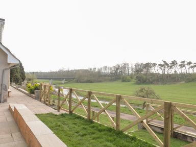 A patio with stone tiles and a wooden fence overlooking a grassy area with a river and trees at Whiteadder Bank in Berwick-Upon-Tweed