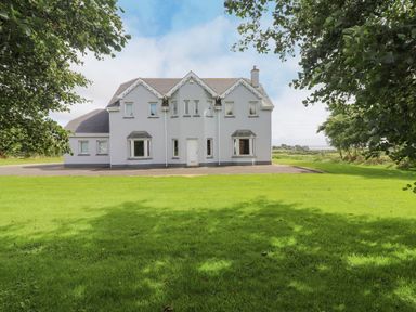 A gray two-story house with multiple windows and a white door surrounded by green lawn and trees at Cloughoge House in Kilrush County Clare