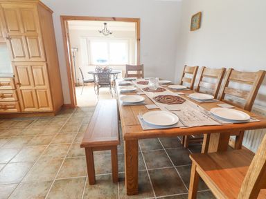 A dining area with wooden table and chairs set with plates and cutlery next to a kitchen with wooden cabinets and a second dining table in the background at Cloughoge House in Kilrush County Clare