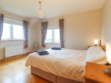 A bedroom with a double bed two windows floral curtains wooden floors and bedside tables with lamps at Cloughoge House in Kilrush County Clare