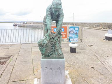 A bronze statue of a person bending over handling a net on a waterfront with a pier and informational signs in the background at Cloughoge House in Kilrush County Clare