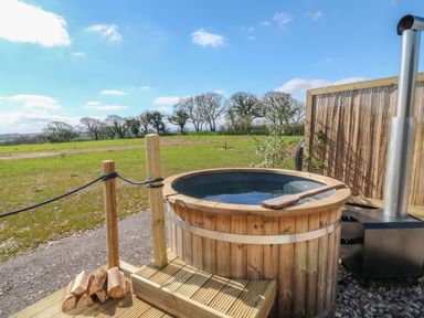 An outdoor hot tub made of wood with a metal stove and a wooden paddle on a deck with grass and trees in the background at Buzzards in Morchard Bishop