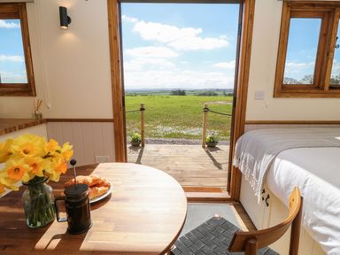 A small dining area with a wooden table and chair next to a bed and an open door leading to a green field at Buzzards in Morchard Bishop