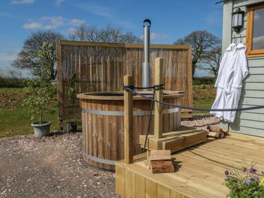 A wooden hot tub with chimney on a deck next to two white robes hanging outside a building at Buzzards in Morchard Bishop