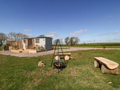 A small wooden cabin with a deck and outdoor furniture next to a fire pit and wooden bench on grass at Buzzards in Morchard Bishop