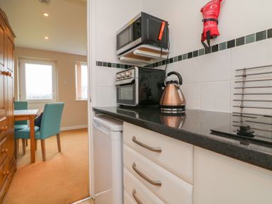 A kitchen counter with a kettle toaster microwave and fridge next to a dining area with blue chairs and wooden table at Sky Pocket in Bude