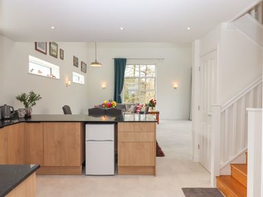 A kitchen with wooden cabinets a small white fridge and a view into a living area with a large window and a staircase on the right at Narracott Down in South Molton