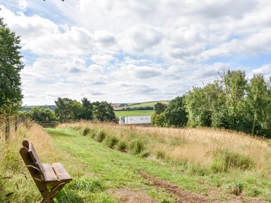 A wooden bench along a grassy path with trees and a house in the distance at Narracott Down in South Molton