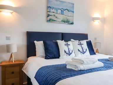 A bedroom with a bed featuring navy and white pillows with anchor designs bedside tables with lamps and a painting of beach huts above the bed at Strawberry Bank - Nuthatch in Pyworthy near Bridgerule