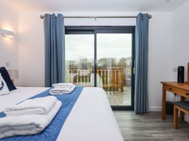 A bedroom with a bed, blue curtains, a wooden desk with a chair, and sliding glass doors leading to a patio at Strawberry Bank - Nuthatch in Pyworthy near Bridgerule