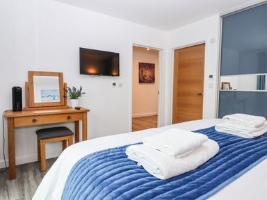 A bedroom with a bed with white towels on a blue runner a wooden dressing table and a wall mounted television at Strawberry Bank - Nuthatch in Pyworthy near Bridgerule