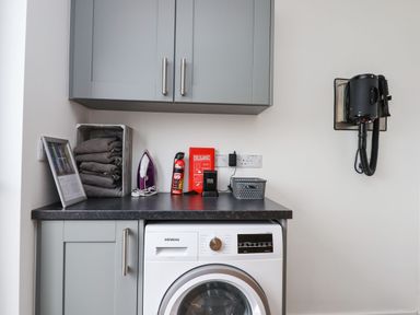 A laundry room with a washing machine under a counter and grey cabinets with an iron and fire safety items at Strawberry Bank - Nuthatch in Pyworthy near Bridgerule