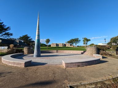 An outdoor circular stone seating area with a tall conical sculpture in the center surrounded by grass trees and buildings at Strawberry Bank - Nuthatch in Pyworthy near Bridgerule