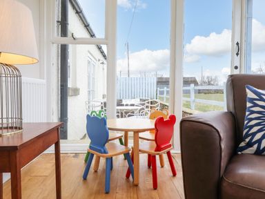 A small wooden table with colorful children's chairs next to a brown sofa and a wooden side table with a lamp by glass doors at Farnelea in West Fleetham near Seahouses