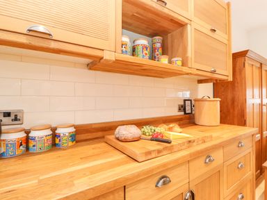 A wooden kitchen counter with jars labeled coffee sugar and tea a cutting board with bread grapes tomatoes and cheese and wooden cabinets at Farnelea West Fleetham near Seahouses