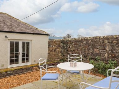 An outdoor patio area with a white round table and white chairs with blue cushions near a stone wall and a building with double glass doors at Farnelea in West Fleetham near Seahouses