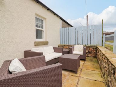 An outdoor patio area with brown wicker furniture and white cushions next to a white wall and blue fence at Farnelea in West Fleetham near Seahouses