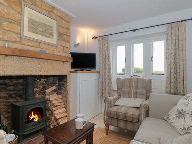 A sitting room with a wood burning stove and stacked firewood a plaid armchair a sofa windows with curtains and a small table with two mugs at Scrambles Cottage Stoke-Sub-Hamdon Somerset
