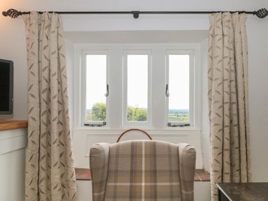 A window with three panes flanked by curtains and a plaid armchair facing the window at Scrambles Cottage in Stoke-Sub-Hamdon Somerset