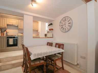 A dining area with a wooden table and chairs next to a radiator and a large wall clock with a kitchen visible in the background at Scrambles Cottage in Stoke-Sub-Hamdon Somerset
