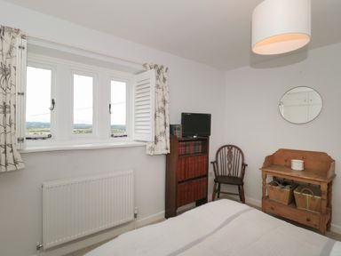 A bedroom with a window with curtains a wooden chair a wooden shelf with baskets and a small TV at Scrambles Cottage in Stoke-Sub-Hamdon Somerset