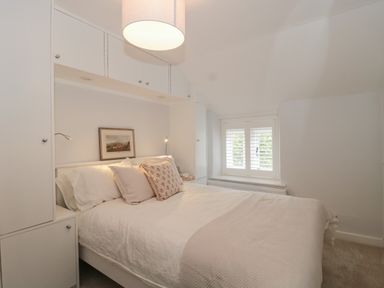 A bedroom with a bed and pillows under built in cabinets and a window with shutters at Scrambles Cottage in Stoke-Sub-Hamdon Somerset