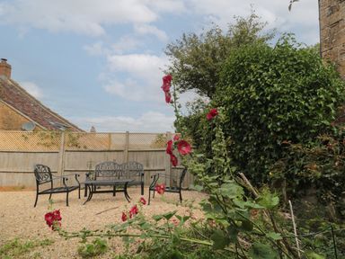 An outdoor seating area with metal chairs and a table on gravel surrounded by flowers and bushes at Scrambles Cottage in Stoke-Sub-Hamdon Somerset