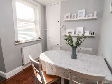 A dining table with four chairs and a vase with leaves on the table in a room with framed photos on shelves at 4 Percy Street in Amble