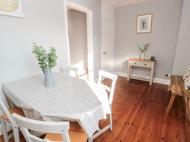A dining area with a table covered by a polka dot tablecloth and white chairs next to a small console table with decorations and a wooden bench on a wooden floor at 4 Percy Street in Amble