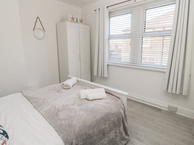 A bedroom with a bed covered with a gray blanket and rolled towels white wardrobe round mirror and window with white blinds and curtains at 4 Percy Street in Amble