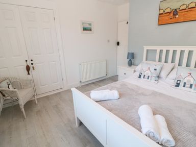 A bedroom with a white bed, pillows, rolled towels, nightstand with lamp, wicker chair, closet doors, and wall art at 4 Percy Street in Amble